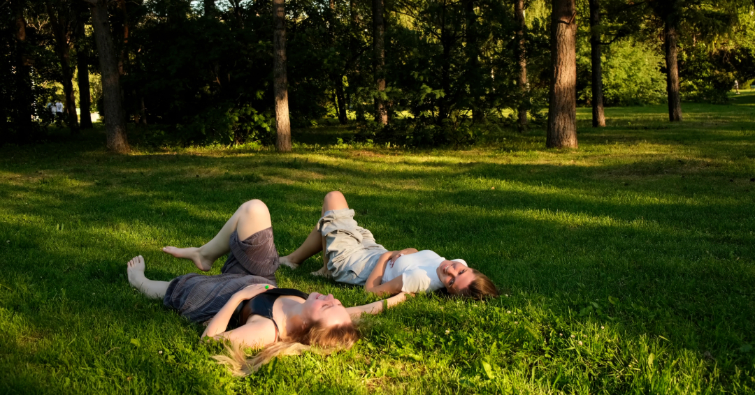 Two girls laying in green grass at golden hour, laughing with each other.