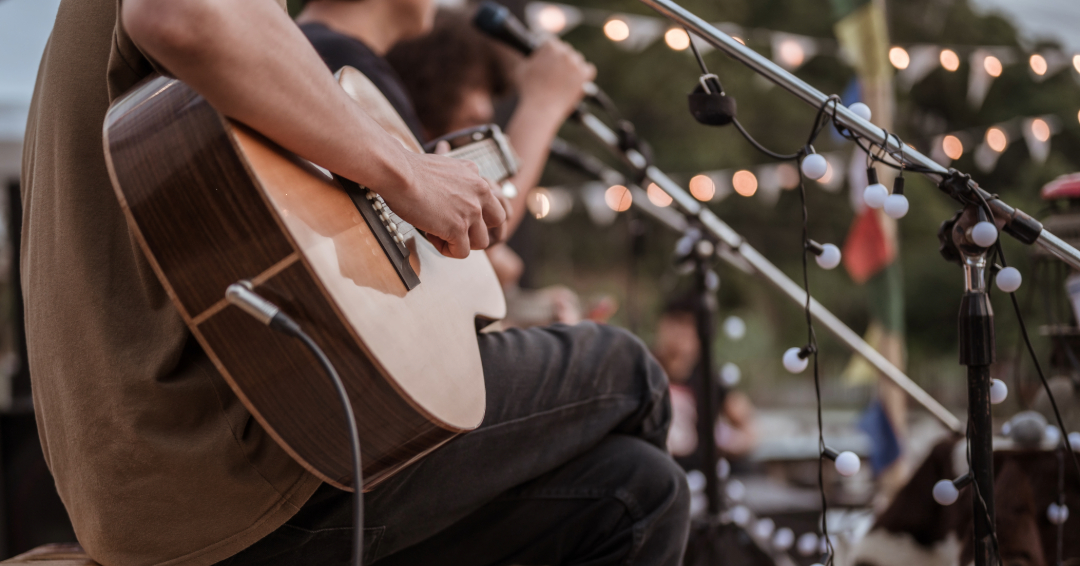 A performer strums an acoustic guitar on stage, outdoors.