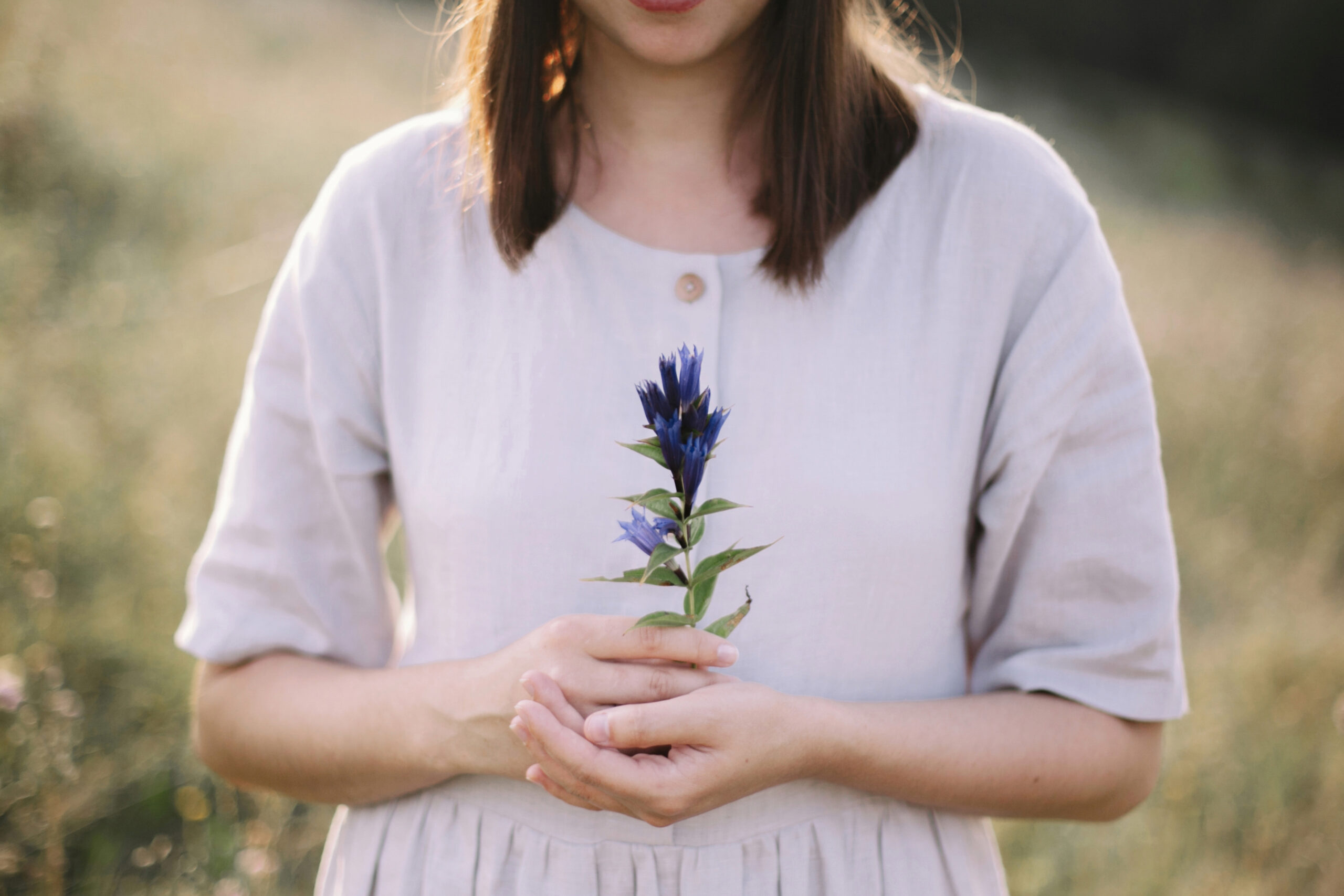 A woman wearing a neutral dress holds a blue flower in her hands.