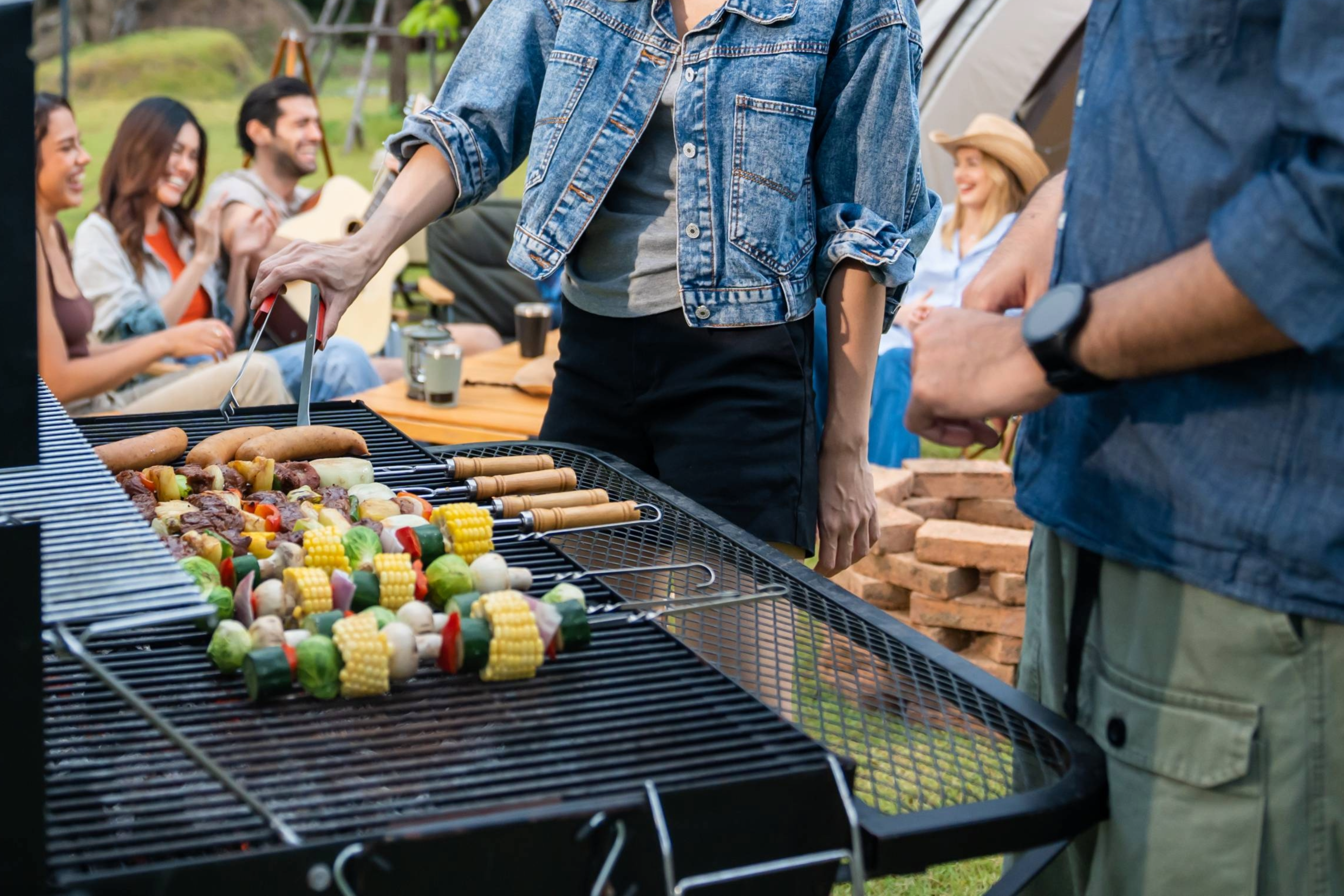 A group of friends is having a cookout, grilling brats and vegetable skewers.