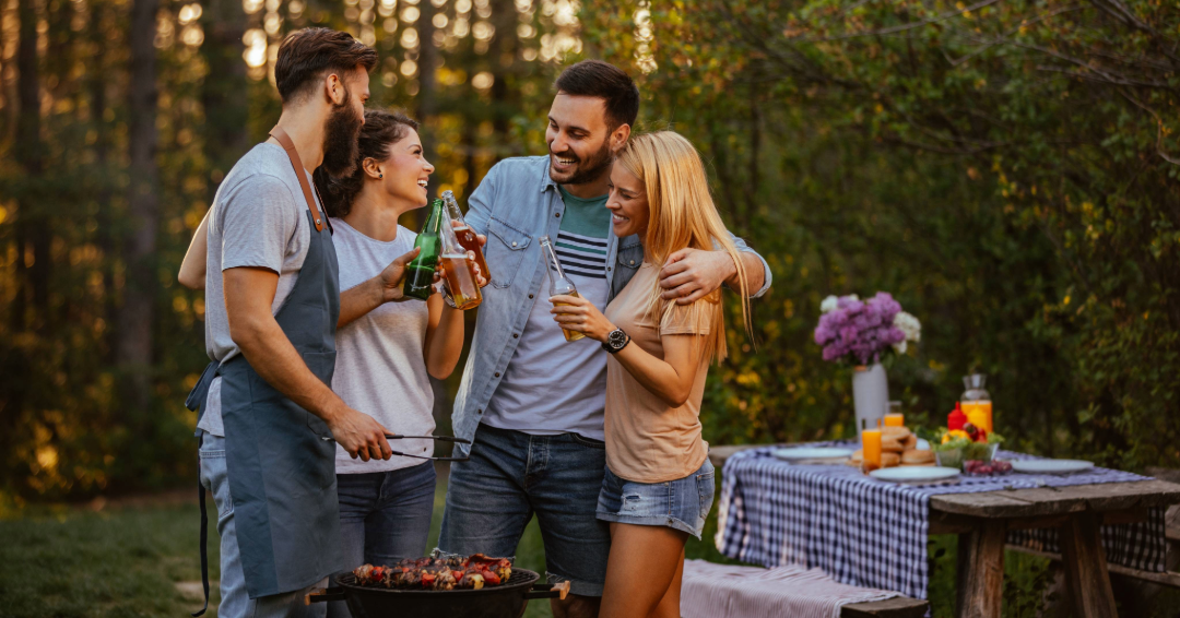 Friends, gathered around a grill, cheers and laugh at an evening summertime cookout. 