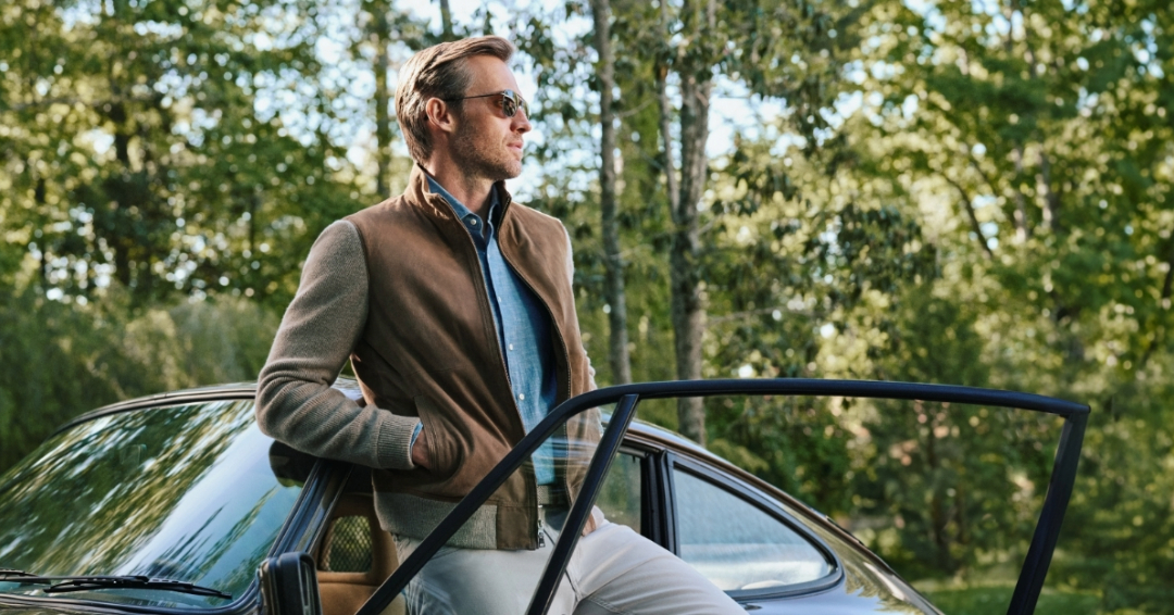 Menswear in Franklin and Brentwood, TN, featuring a man leaning against a car door in a wooded outdoor setting while wearing a brown jacket, blue shirt, sunglasses, and tailored casual layers.